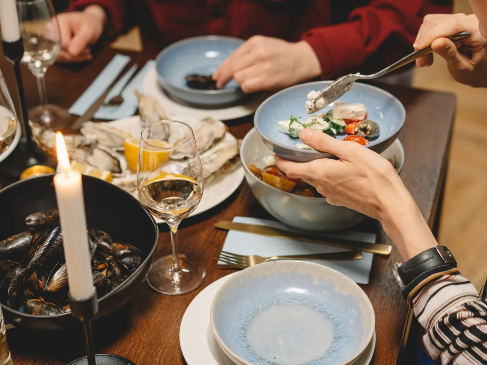 Hände, die Meeresfrüchtegerichte an einem mit Wein Gläser gedeckten, kerzenbeleuchteten Holztisch teilen